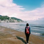 Back view of a woman walking along a serene coastal beach with a backpack.
