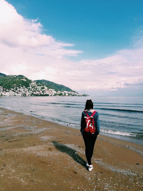 Back view of a woman walking along a serene coastal beach with a backpack.