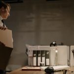 Businessman stands at desk packing documents in a dimly lit office setting.